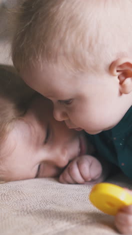 funny girl swings legs lying on large cosy bed and hugging little brother with yellow toy circle in light room at home