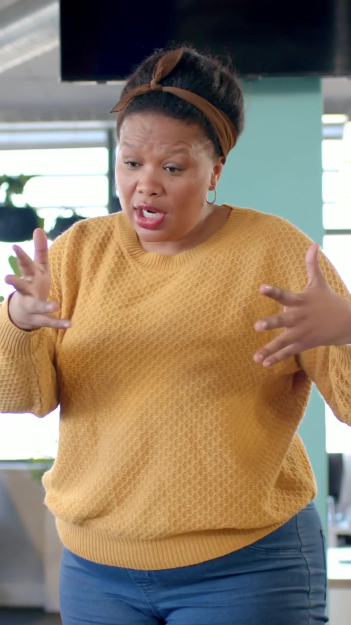 Vertical video: Starting to talk, woman in sweater pointing and clarifying in office lounge with TV
