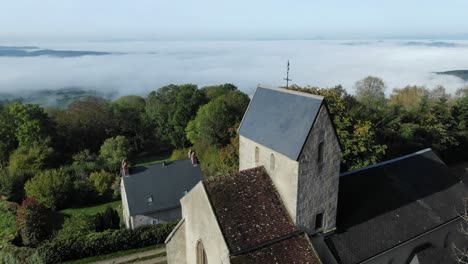 drone flying over church of saint-roch in uchon with fog shrouded valley in background, saone-et-loire department in france