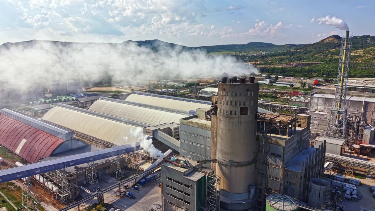Factory in operation with smoke and machinery under a cloudy sky near a hillside landscape