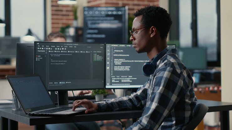 Portrait of african american developer using laptop to write code sitting at desk with multiple screens parsing algorithm