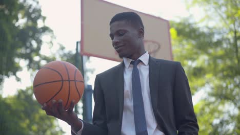 portrait of elegant african american businessman posing with ball on basketball court. middle shot of confident young man in formal suit keen on sport. life balance concept.