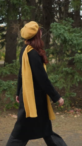 estudiante emocionada con largo cabello rojo ondulado con un abrigo negro, boina amarilla, y bufanda a juego, caminando a través de un exuberante parque verde con colorido follaje de otoño, felizmente levantando la mano a la cabeza
