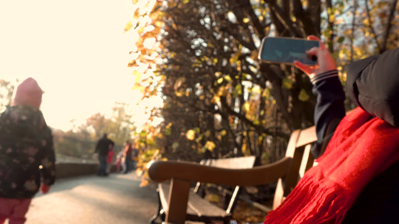 Beschnittene Hand einer jungen Frau, die auf einer Bank sitzt und mit ihrem Smartphone Fotos macht, während Menschen auf dem Weg im Park Oliwski in Danzig, Polen, vorbeigehen