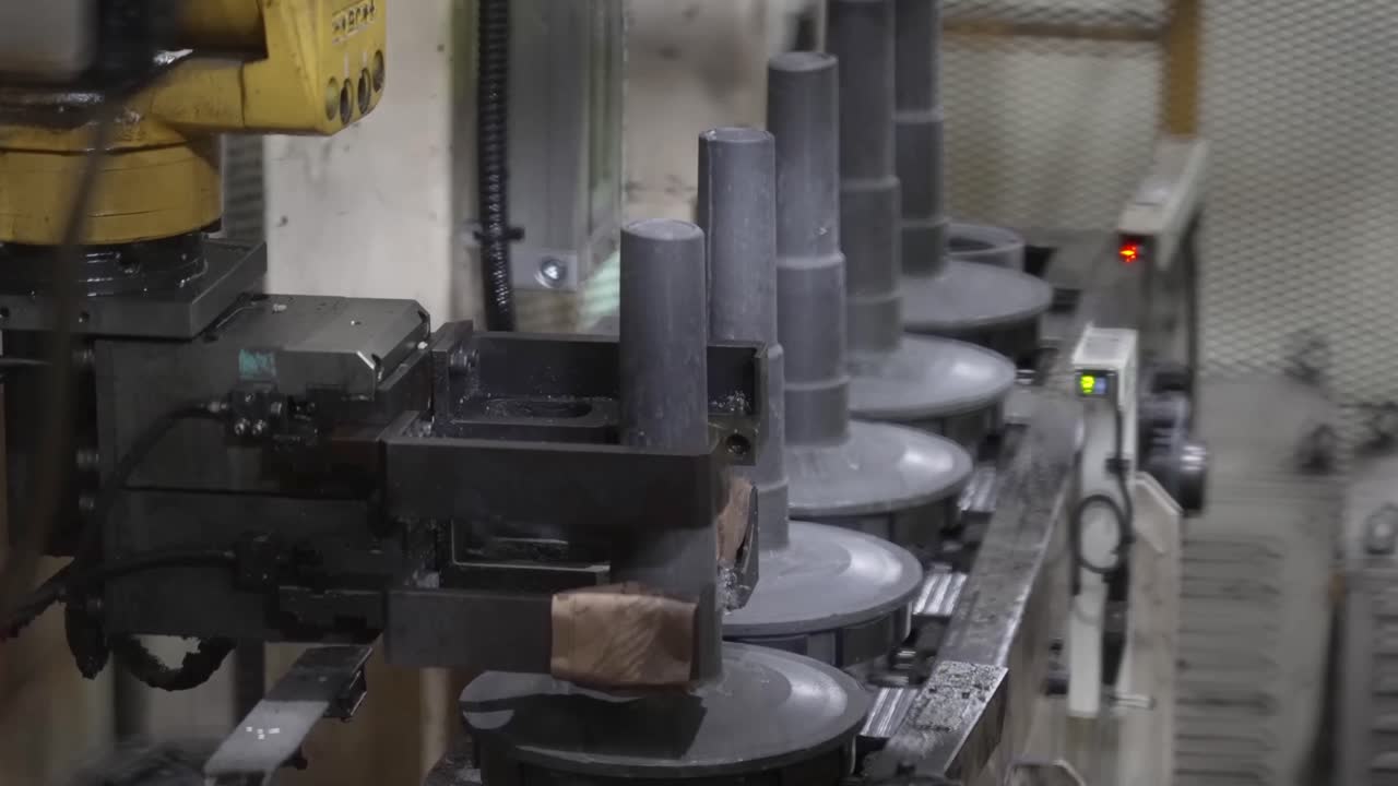Dönen bir montaj istasyonunda dikey silindirik metal iş parçalarını işleyen robotik bir kolun yer aldığı otomatik üretim hattı.