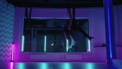 A-woman-does-yoga-and-stretching-on-a-hanging-hammock-in-neon-light.-A-woman-is-flying-in-a-hammock-in-the-studio-doing-stretching-and-body-exercises.-Exercises-in-the-air