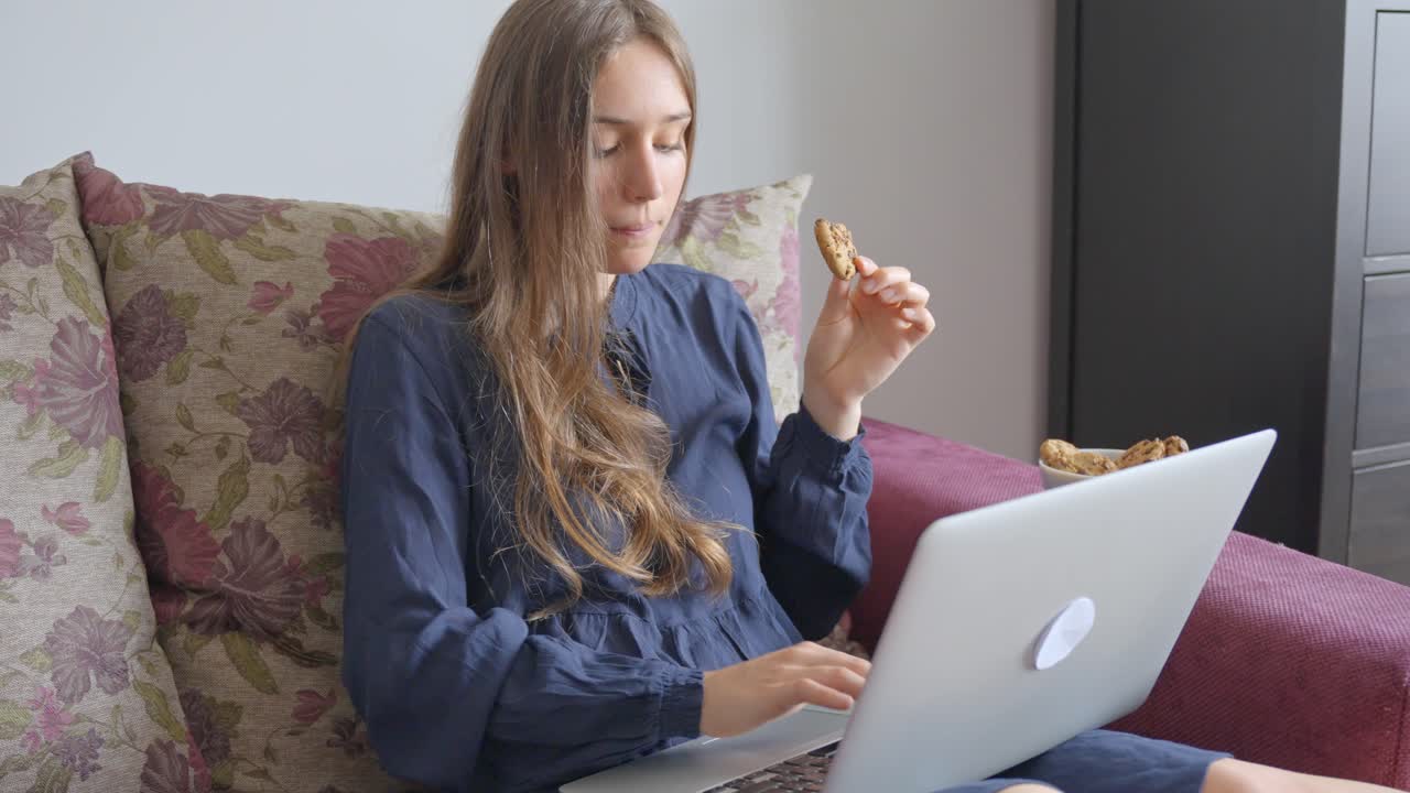 Young woman working from home, sitting on the sofa with a laptop and snacking on cookies, in Slow Motion