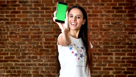chica linda usando el teléfono inteligente, deslizando, mostrando la pantalla en la cámara, como el letrero, la llave de croma, la pared de ladrillo en el fondo, el retrato