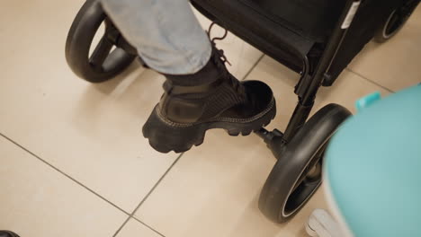 leg view of shopper in black boots and denim trousers testing stroller brake lever with foot, then rolling stroller back and forth on tiled floor in lit baby gear aisle evaluating wheel quality