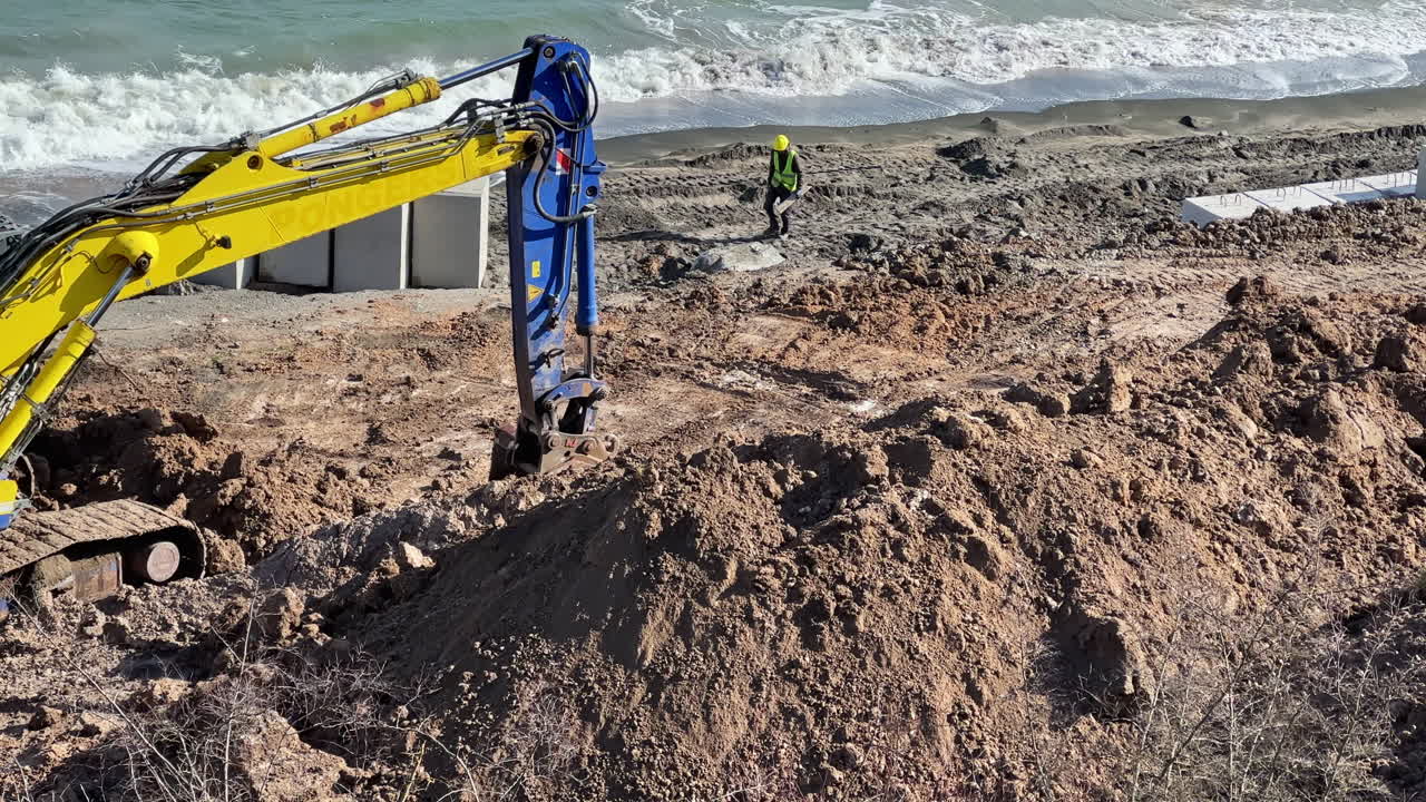 Lavori di costruzione in un'area costiera con macchinari pesanti che movimentano terra vicino all'oceano durante il giorno.