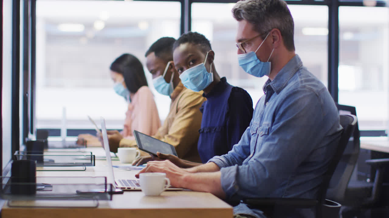 현대적인 사무실에서 책상에 앉아 일하는 사무실 동료들이 얼굴 마스크를 착용하고 있습니다.