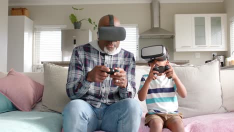 african american grandfather and grandson sitting on sofa, having fun, using vr headset, slow motion