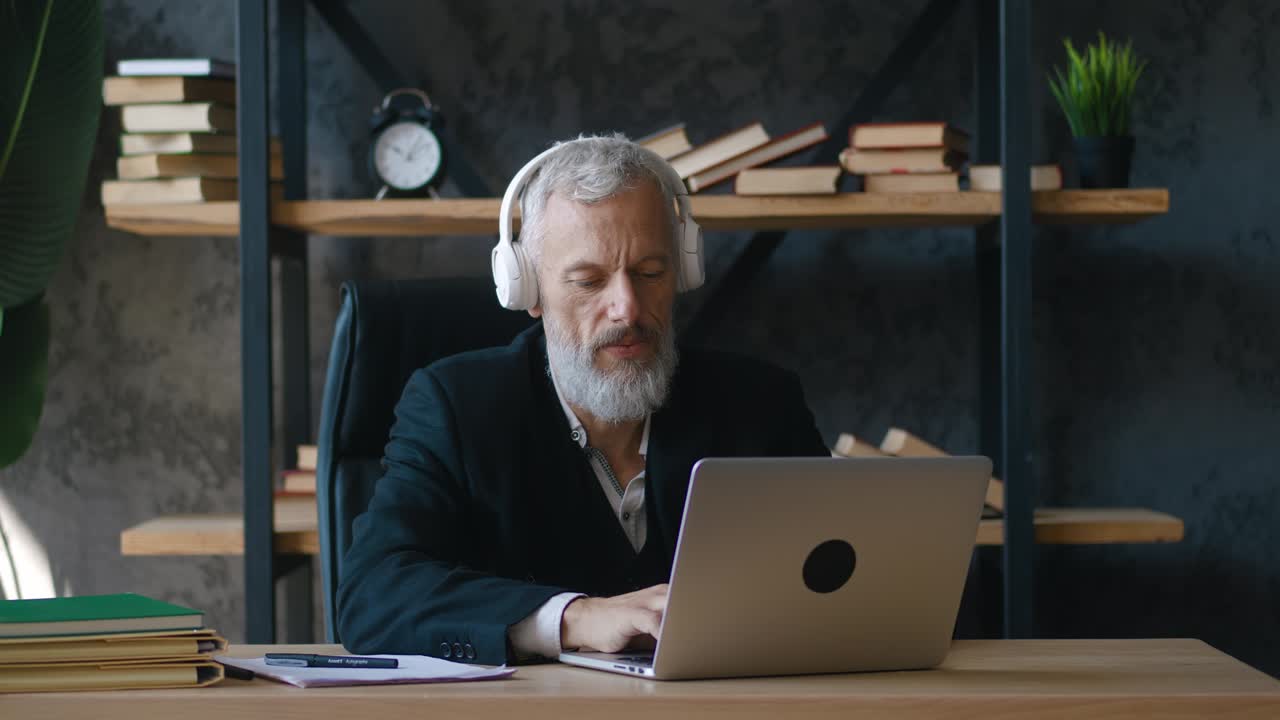 Happy bearded mature businessman listening music in headphones while working at laptop computer at office