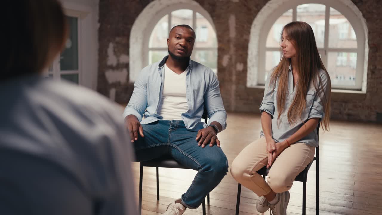 A fit man with Black skin in a blue shirt and a girl with long hair communicate with a psychologist during group therapy