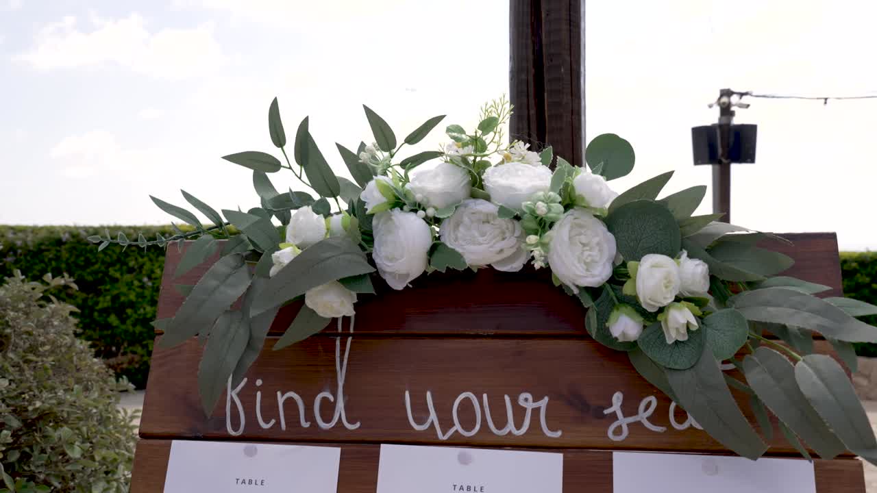 An outdoor wedding setup is showcased with a wooden sign featuring floral accents. The sign invites guests to find their seats while the sun shines and greenery surrounds the area