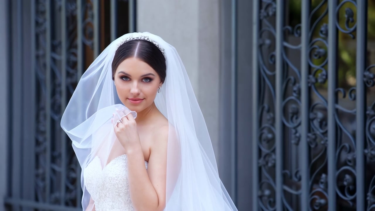 Una sposa in abito di pizzo bianco e lungo velo sta accanto a un cancello di ferro ornato alla luce del giorno, con lo sguardo rivolto di lato e un'espressione calma ed elegante.