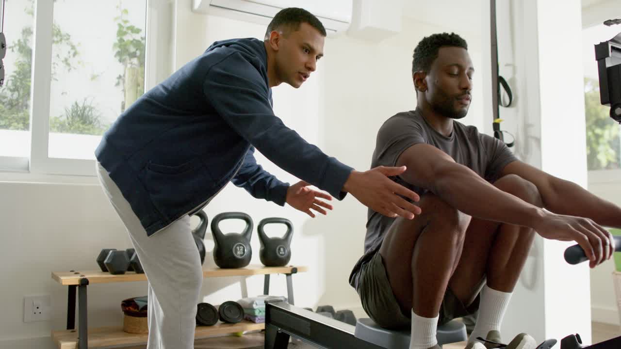 Trainer guiding mid adult African American man rowing on rower for posture correction in home gym