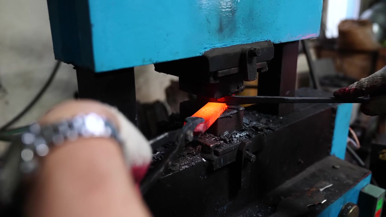 Gloved hands guiding a red hot steel bar under the die of a power hammer during the forging of a blade