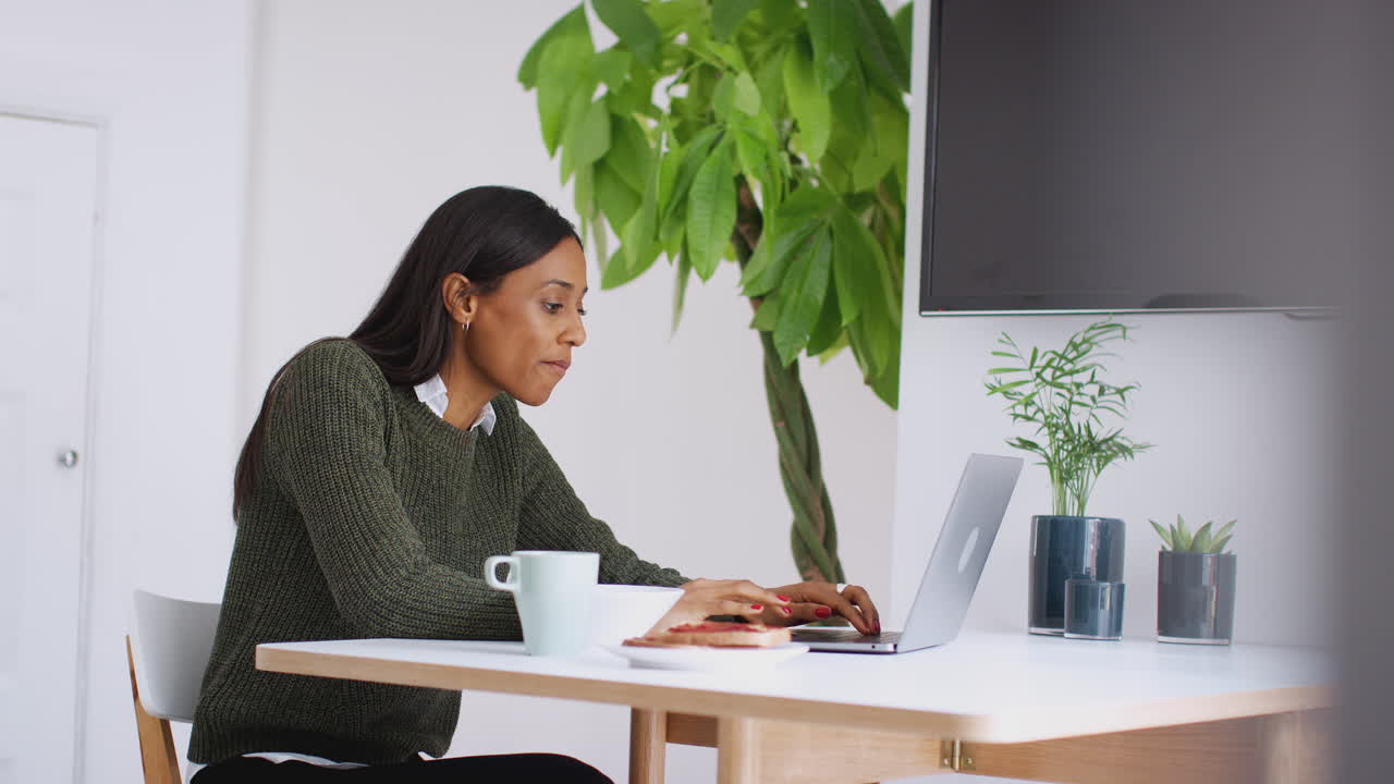 Businesswoman At Home Eating Breakfast Looking At Laptop Before Leaving For Work