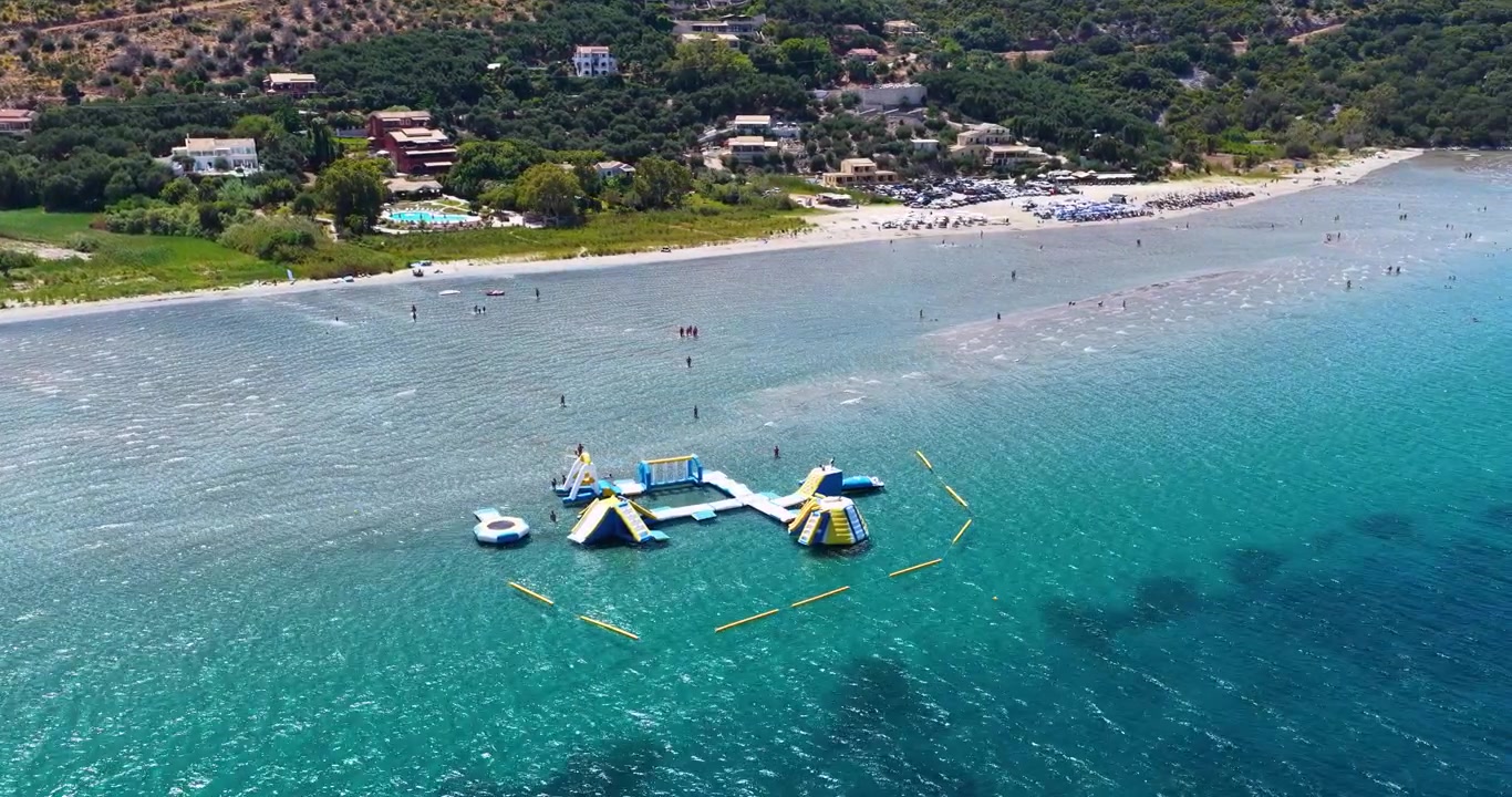 Yunanistan Korfu'daki Apraos Kalamaki plajının kumlu kıyı şeridinin ve sığ mavi sularının, şişme bir su parkı ve yüzücülerle birlikte yüksek açılı drone çekimi