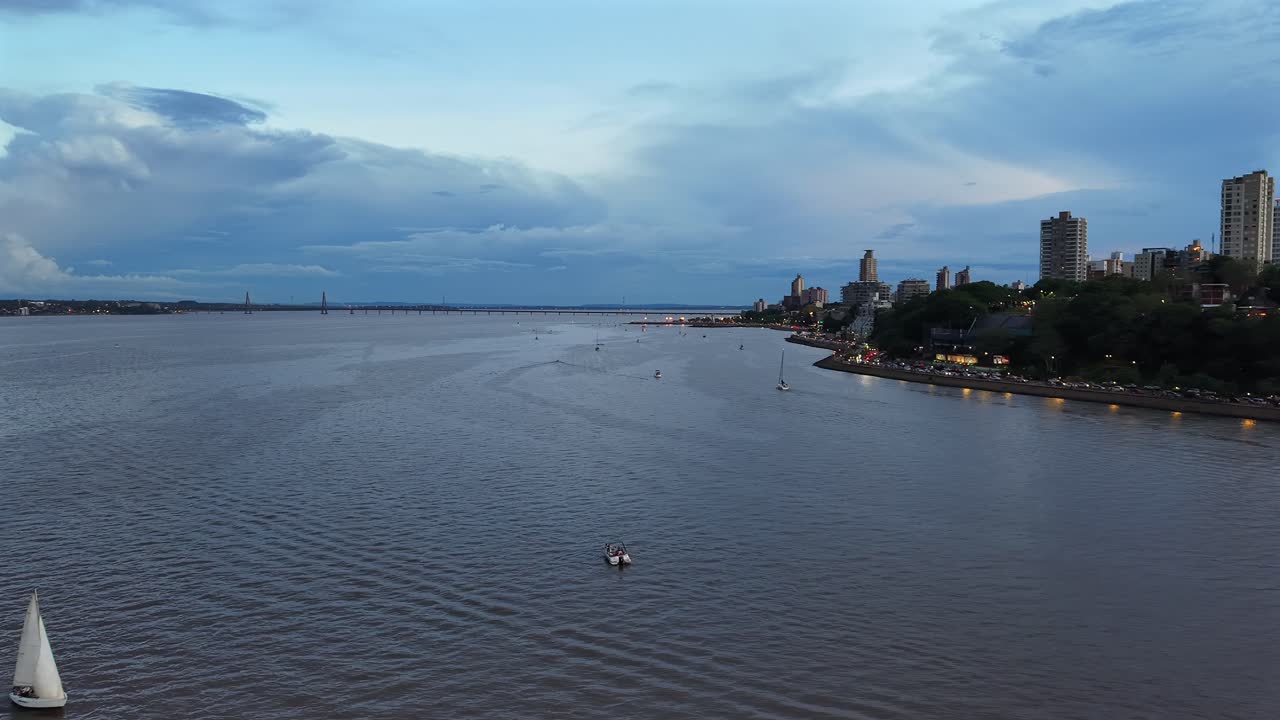 Clip aerea lenta che si muove sulle acque del fiume Paraná a Posadas, Misiones. Il clip cattura l'atmosfera mattutina mentre il cielo si rasserena dopo una tempesta, con nuvole, barche a vela sull'acqua e la città sulla destra.