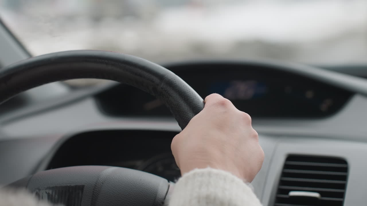 Close up hand wearing white sleeve holding steering wheel while driving car with blurred dashboard in background and snowy landscape outside window, focus on hand and steering wheel