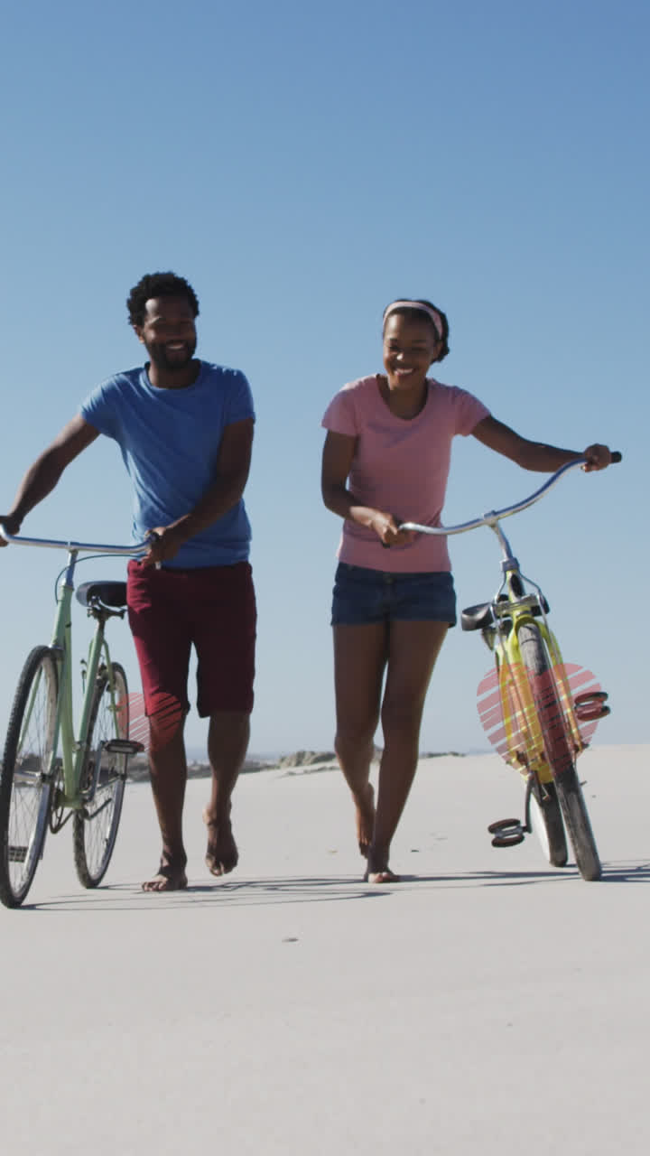 Animation of hearts over happy african american couple walking with bikes on beach