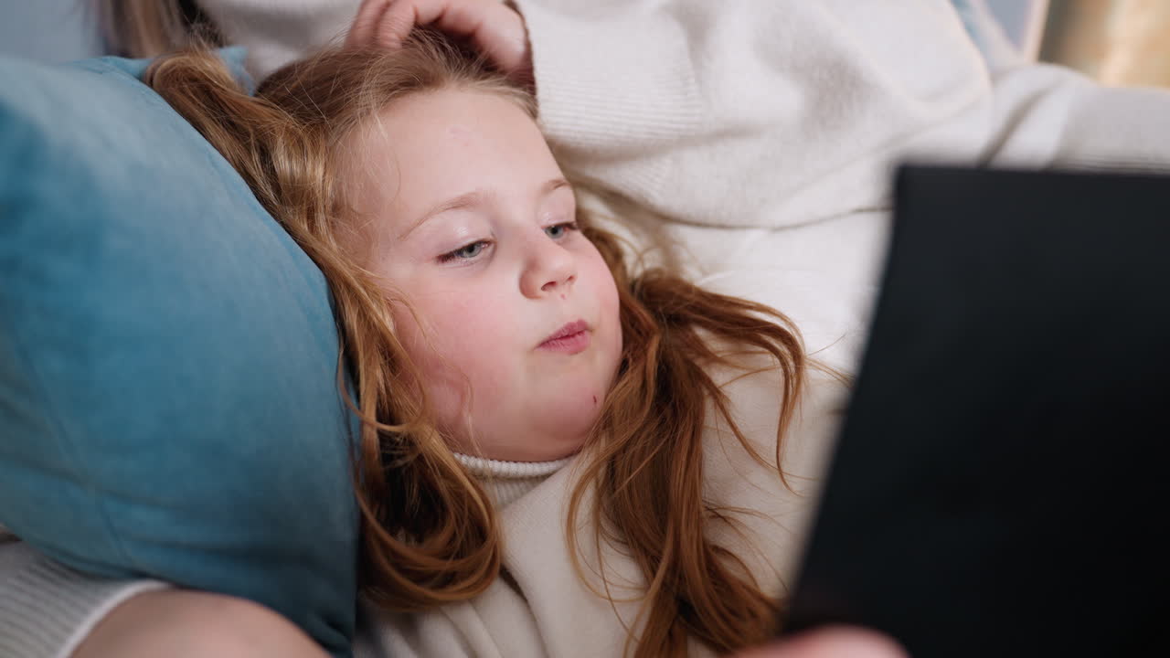 Bambina sdraiata sul letto che tiene in mano un libro, labbra leggermente dischiuse mentre legge ad alta voce con attenzione e coinvolgimento, atmosfera accogliente con abiti caldi, che evidenzia l'apprendimento infantile, momento di tranquillità domestica