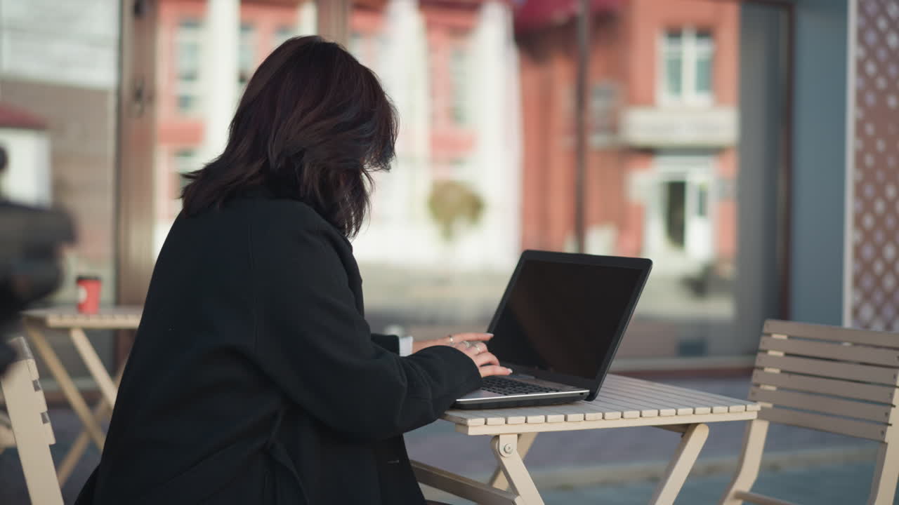 Close up of woman working on laptop outdoors with hair covering her face, coffee cup on table beside her, and blurred view of urban building and street in background