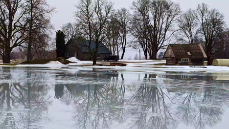 Paisaje-Invernal-De-Charco-De-Deshielo,-Casas-Rústicas-En-Reflexión,-Vista-Increíble-Del-Campo-De-Letonia