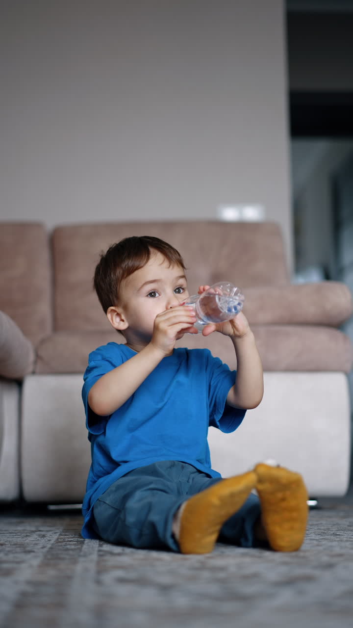 Beautiful Caucasian toddler sits on the floor at home. Kid drinks water from bottle. Black cat approaches. Vertical video.