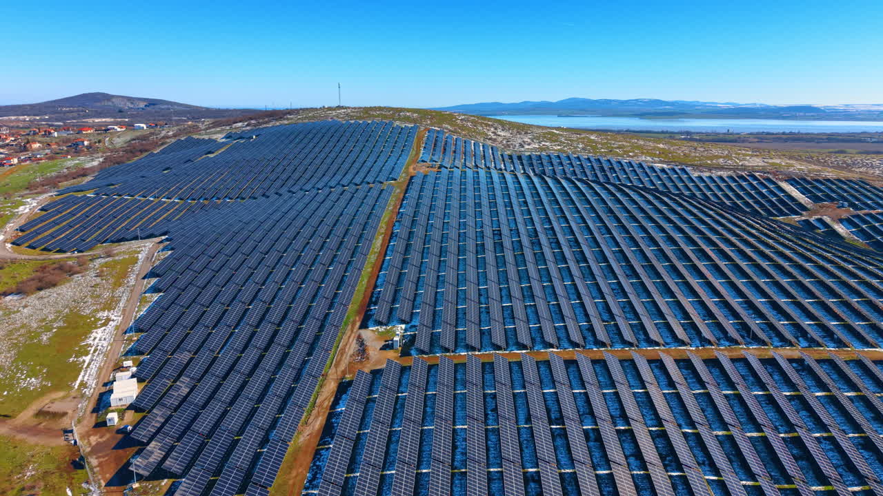 Luftfoto af stor solcellepark på en bjergskråning. Solcellepaneler på en grøn bakke nær en sø og landsby under klar blå himmel