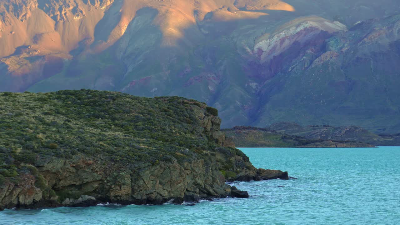 Steady shot of Lake Belgrano's turquoise shoreline in Patagonia with gentle waves crashing against rocky shore and distant mountains with sunset light. Perito Moreno National Park