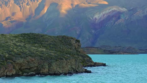 steady shot of lake belgrano's turquoise shoreline in patagonia with gentle waves crashing against rocky shore and distant mountains with sunset light. perito moreno national park