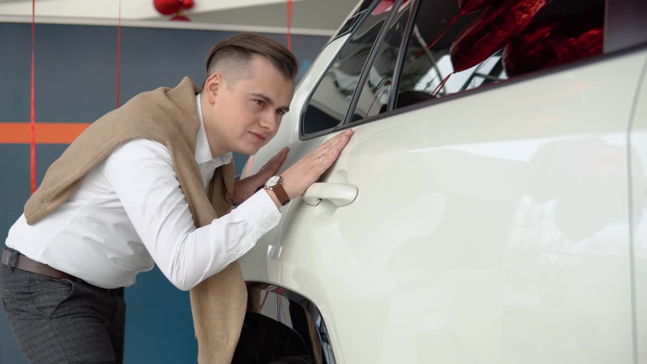 Guy touches the side door of a shiny white car. Young man looking carefully at car in car dealership
