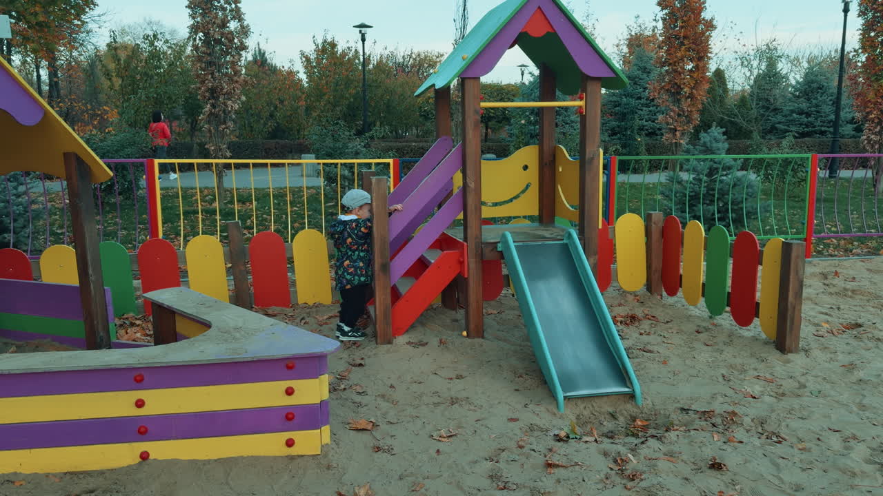 Lovely kid climbs on the slide and taps his feet happily. Baby boy playing on the playground in autumn.