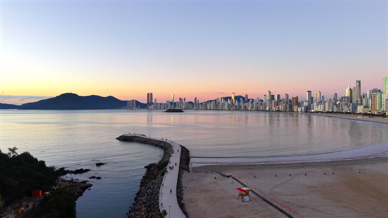 Drone slowly ascends above a curved jetty at sunset, revealing the serene waters of the Gulf of Balneário Camboriú, the distant Ilha das Cabras, and the full skyline bordered by Serra do Mar ridges