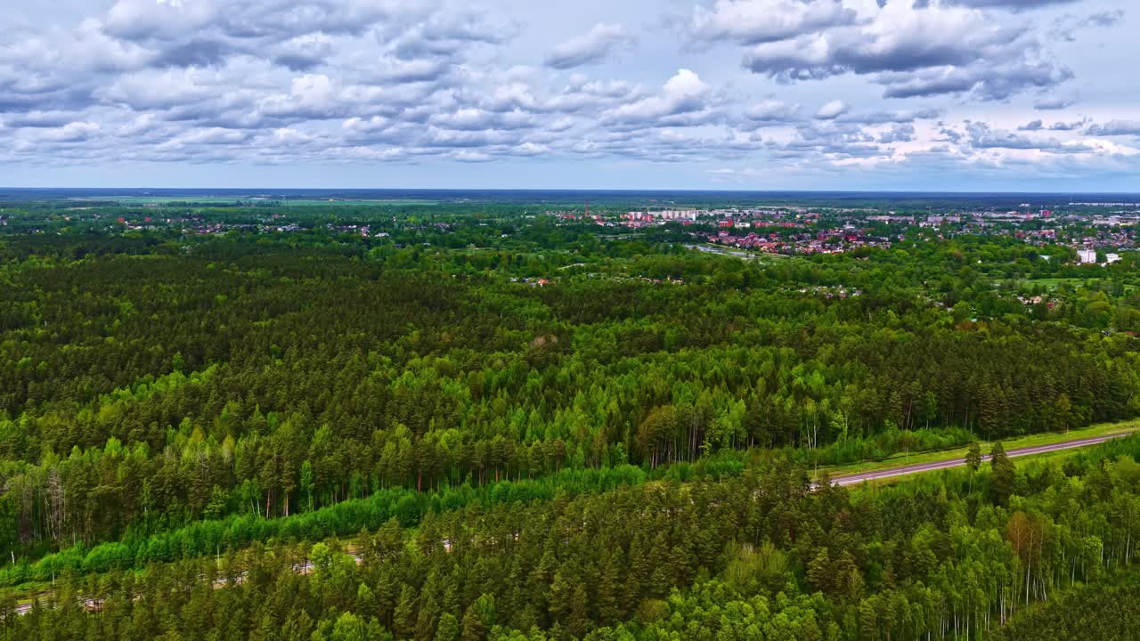 Panoramatický záběr z dronu nad hustými lesy rozprostírajícími se k městu pod dramatickou zataženou oblohou.