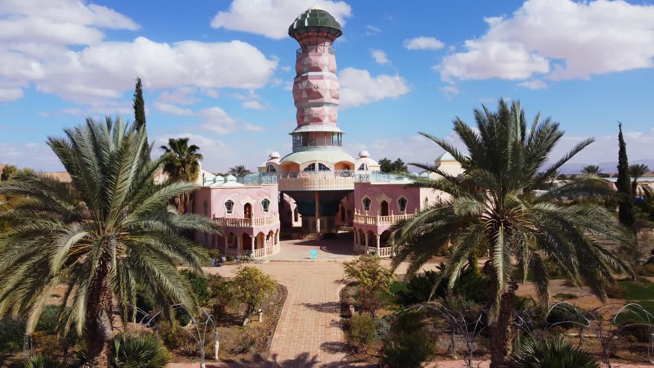 Neot Smadar Art Center building front view with cooling towers under clear blue sky surrounded by palm trees, garden landscapes in Arava desert, Slow motion, aerial tracking shot