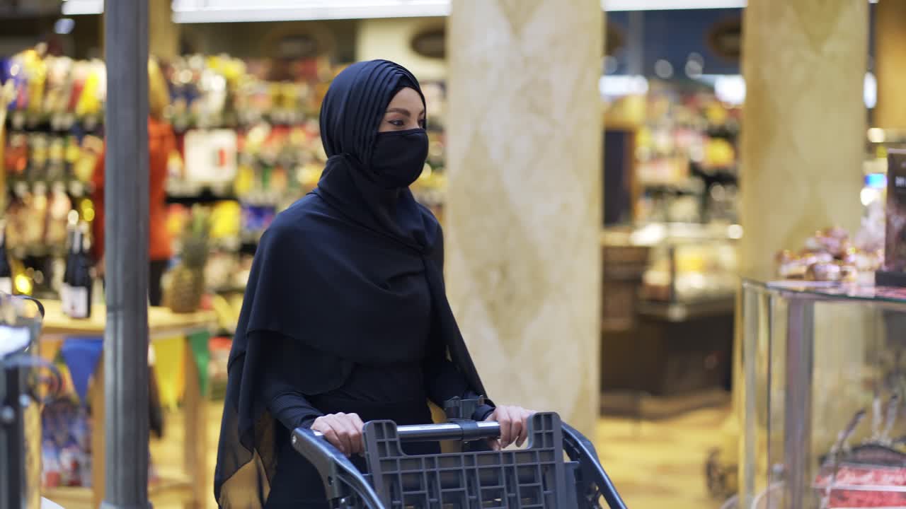 mujer musulmana moderna en hijab haciendo compras en el supermercado eligiendo productos, usando máscara