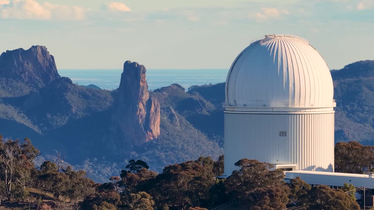 Una grande cupola bianca di un telescopio si trova in cima a un crinale boscoso che si affaccia su cime montuose frastagliate