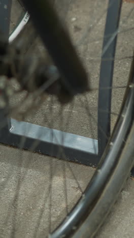 primer plano de una rueda de bicicleta y un soporte de bicicleta metálico en un entorno urbano, capturando detalles de los rayos de las ruedas y el soporte de estacionamiento, otras ruedas de bicicleta visibles en el fondo