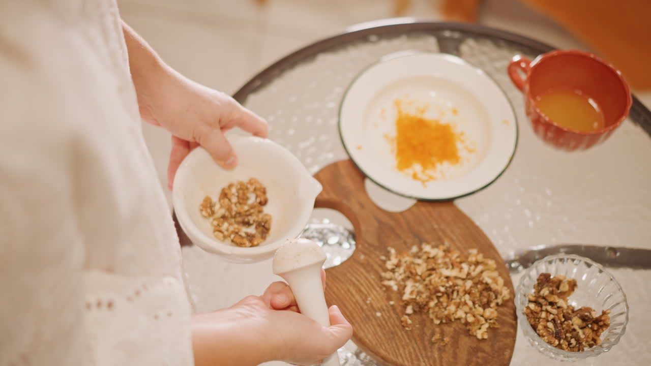 Scatto aereo di cucina con ciotola di noci, mani che mettono noci tritate nella ciotola accanto a scorza di zucca e succo d'arancia, tagliere di legno e cucchiaio dosatore visibili, luce naturale soffusa, costante e metodico.