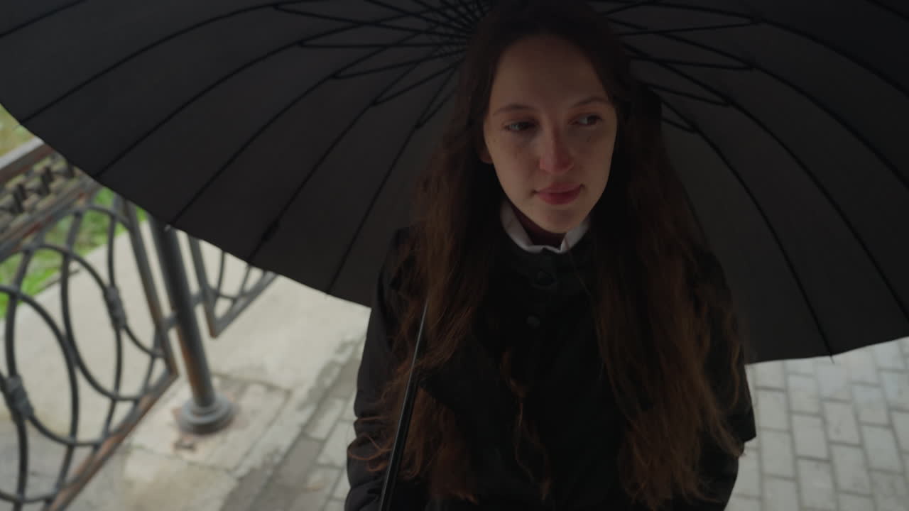Giovane donna in piedi sotto un ombrello nero sul balcone con ringhiera in ferro battuto, capelli lunghi e cappotto scuro, sguardo contemplativo e sottili gocce di pioggia, luce soffusa e ovattata che crea un effetto cinematografico