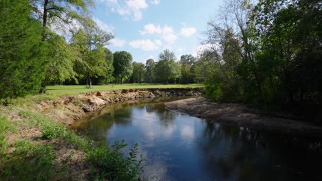 a scenic river flowing through a grassy area with tall, eroded banks and surrounded by lush greenery and trees. the sunny day and clear sky add a peaceful, rural charm to the natural landscape.