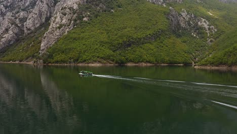 Tour-Boat-Sailing-Near-Mountains,-Creating-a-Picturesque-Scene-with-Calm-Waters-Reflecting-the-Majestic-Mountain-Landscape-in-Albania