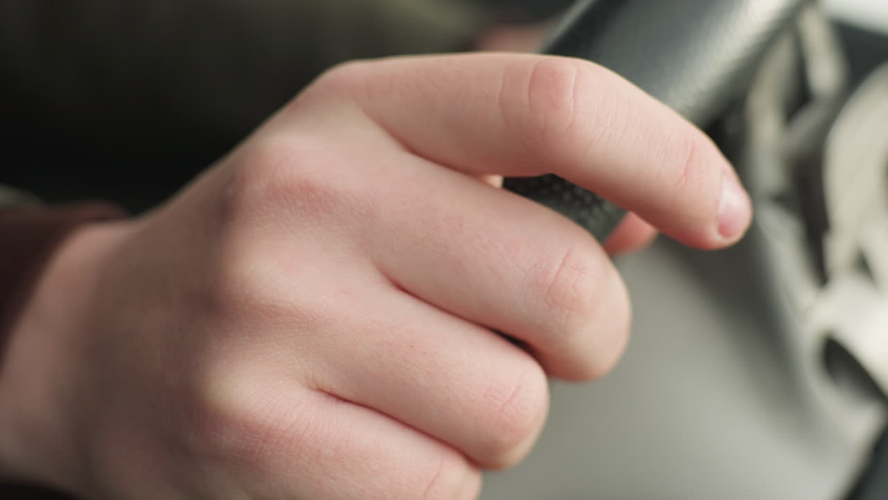 close up of fair skin person gently tapping steering wheel with fingers while holding it firmly, showing casual and calm motion inside car with soft lighting and blurred dashboard area