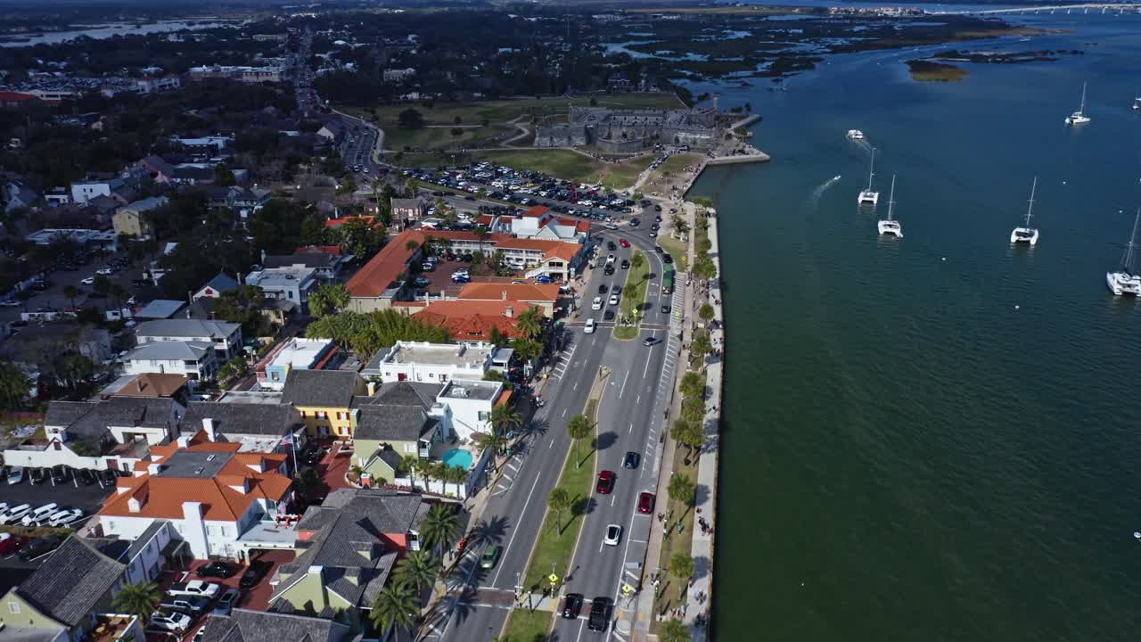 Kystbybillede med en travl vej langs vandet ved siden af en lystbådehavn fyldt med sejlbåde, der ligger for anker i roligt vand, omgivet af bolig- og erhvervsbygninger