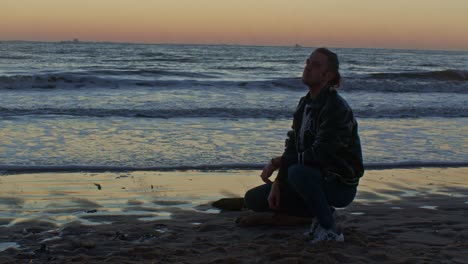 caucasian male man model sits squats on the beach near the sea ocean on the sand shore with sunset and cinematic actor's play