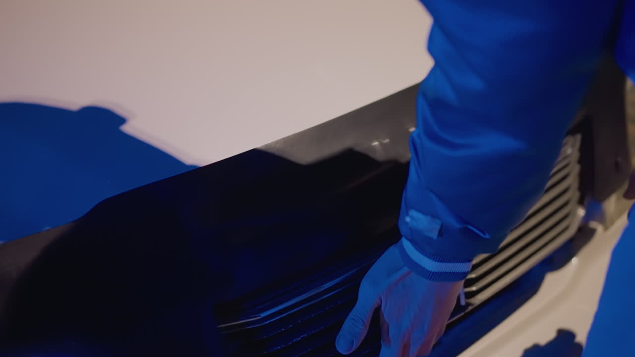 파란색 재킷을 입은 정비공이 극적인 푸른 조명 아래에서 라디에이터 그릴을 손보고 있는 클로즈업 장면입니다. 흰색 자동차 범퍼를 손으로 제거하며 클립 볼트, 플라스틱 트림 및 잠금 장치를 정확하게 움직여 단계별로 작업을 진행합니다.